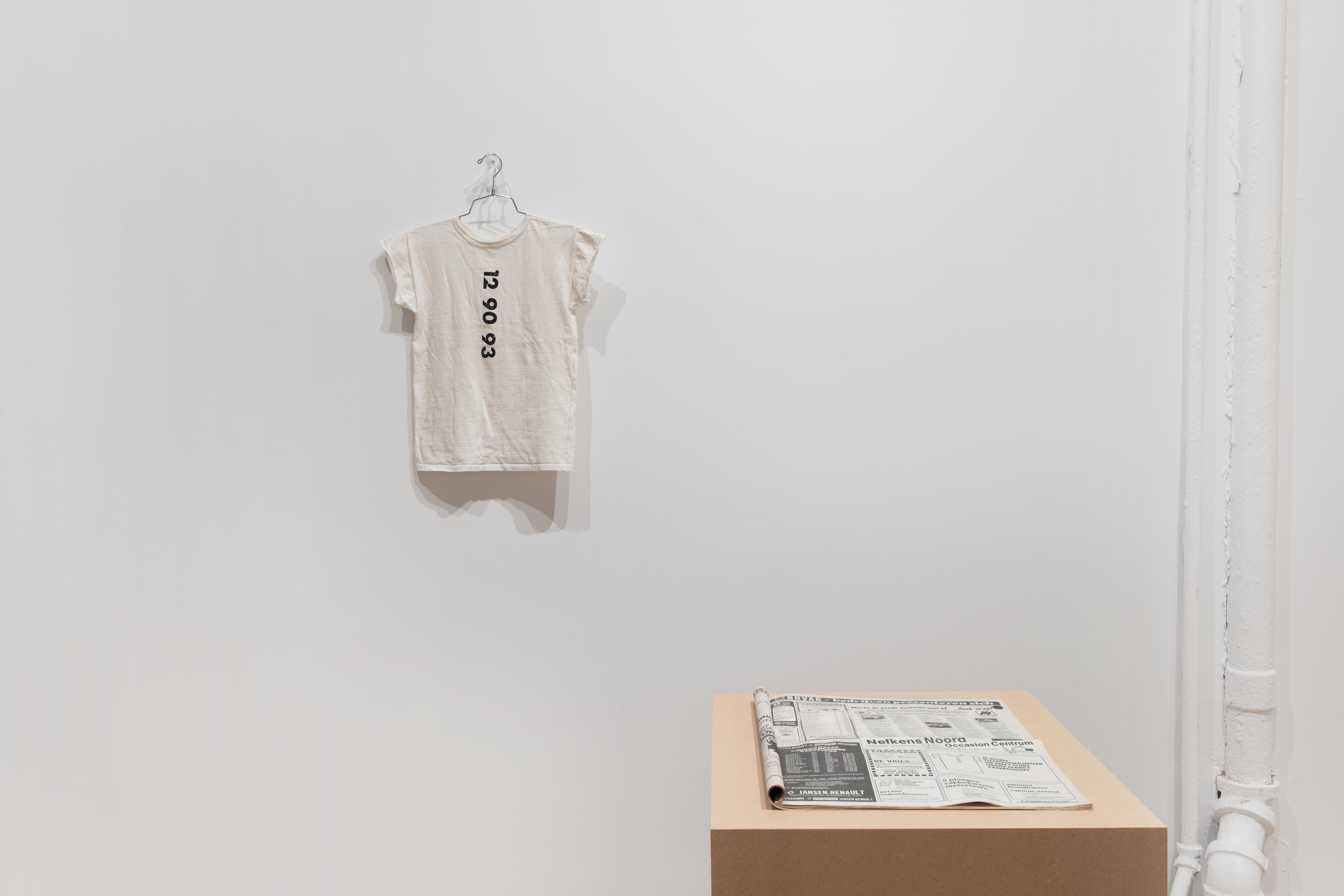 A white T-shirt hangs on a clothes hanger placed on a cylinder of plexi which is affixed to a white gallery wall. Down the middle of the shirt in vertical orientation is a six-digit phone number. In front of the wall and to the right of the shirt is a low plinth made of MDF, with a newspaper placed on top. 