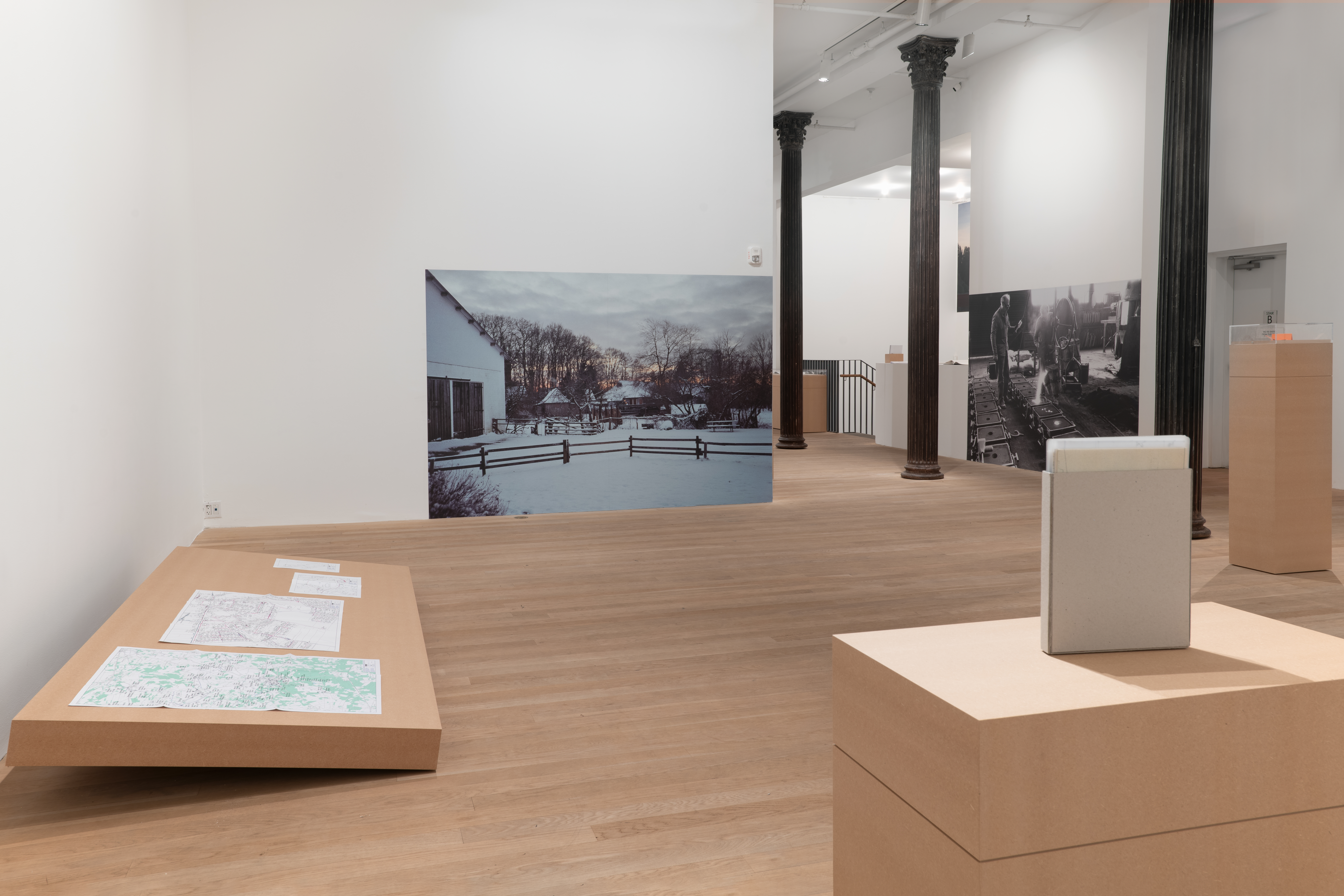 A variety of plinths made of MDF are arranged around a large gallery space. Along the left wall, a low, angled plinth displays a series of annotated utility maps. On the back wall a large photograph is affixed to the wall and depicts the snowy landscape of Neuenkirchen, Germany. 