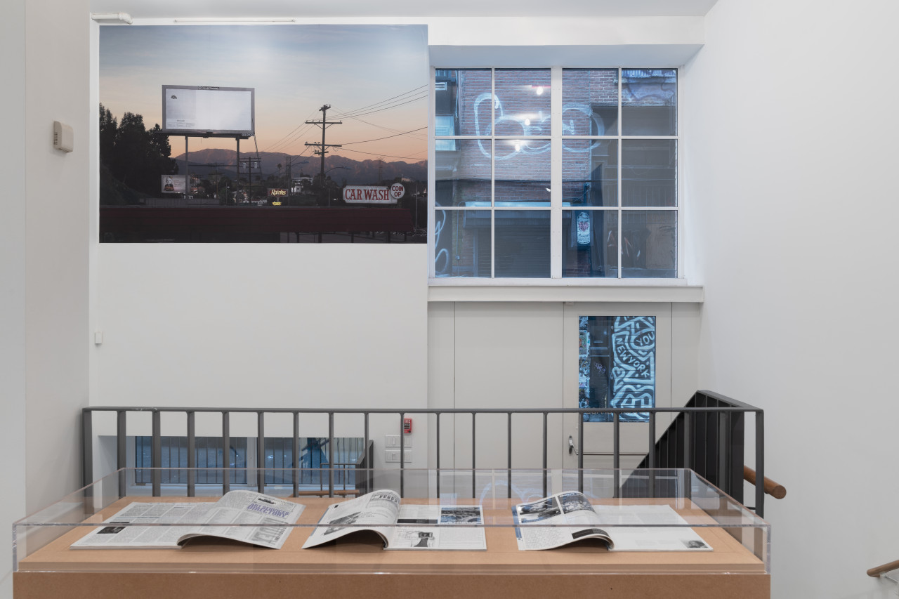 Next to the entrance to a gallery space, along the inside wall, is a large photograph affixed to a wall that depicts a billboard on Glendale Boulevard in Los Angeles. In the foreground is a set of three open magazines displayed on a plinth made of MDF.