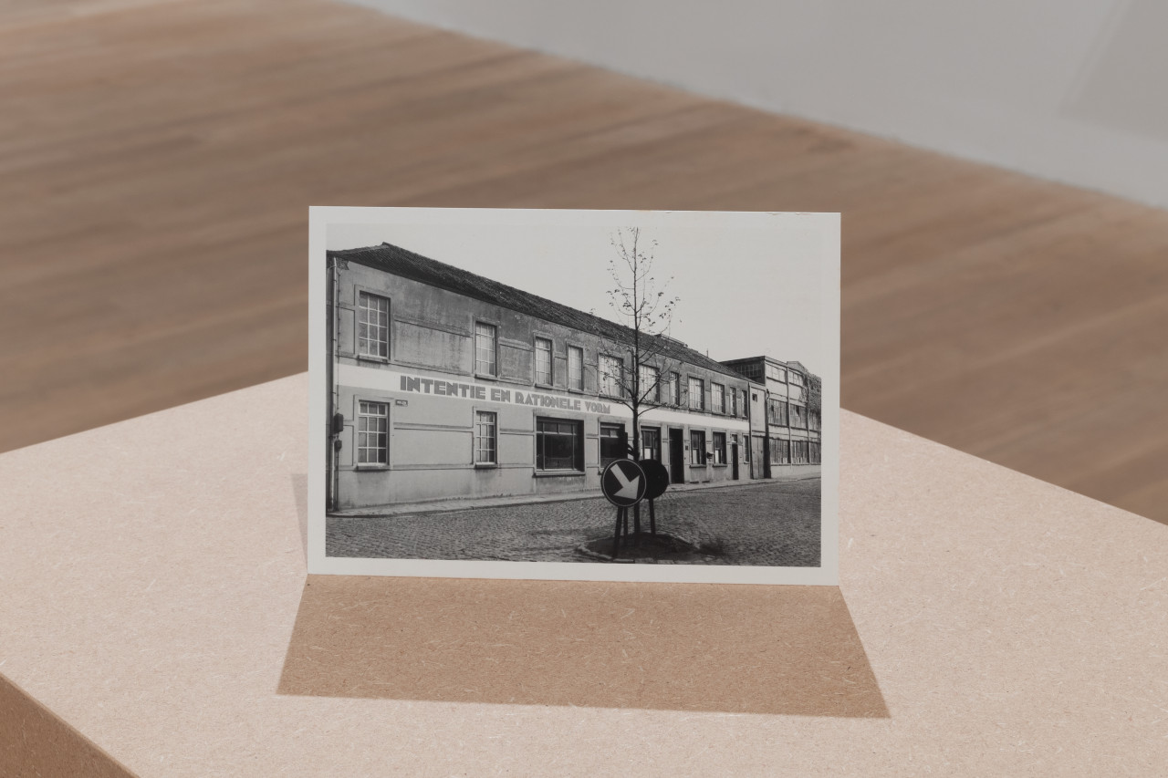 A postcard depicting a black-and-white photograph of the facade of a factory in Belgium sits on a plinth.