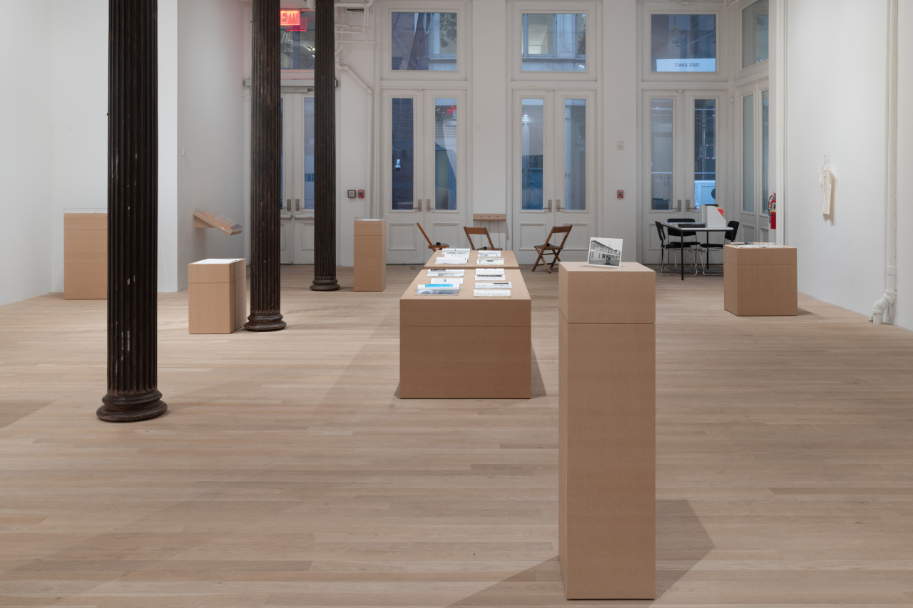A large gallery space houses a variety of plinths made of MDF, atop of which are placed various archival objects. Down the middle of the room are two long, short plinths that display a series of art historical journals. On another tall plinth to the right is a postcard.