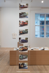 A long piece of plexiglass hangs vertically from the cieling in a gallery. Eight postcards are mounted onto the plexiglass. All of the postcards depict different garbage trucks, shown from the exact same angle. 