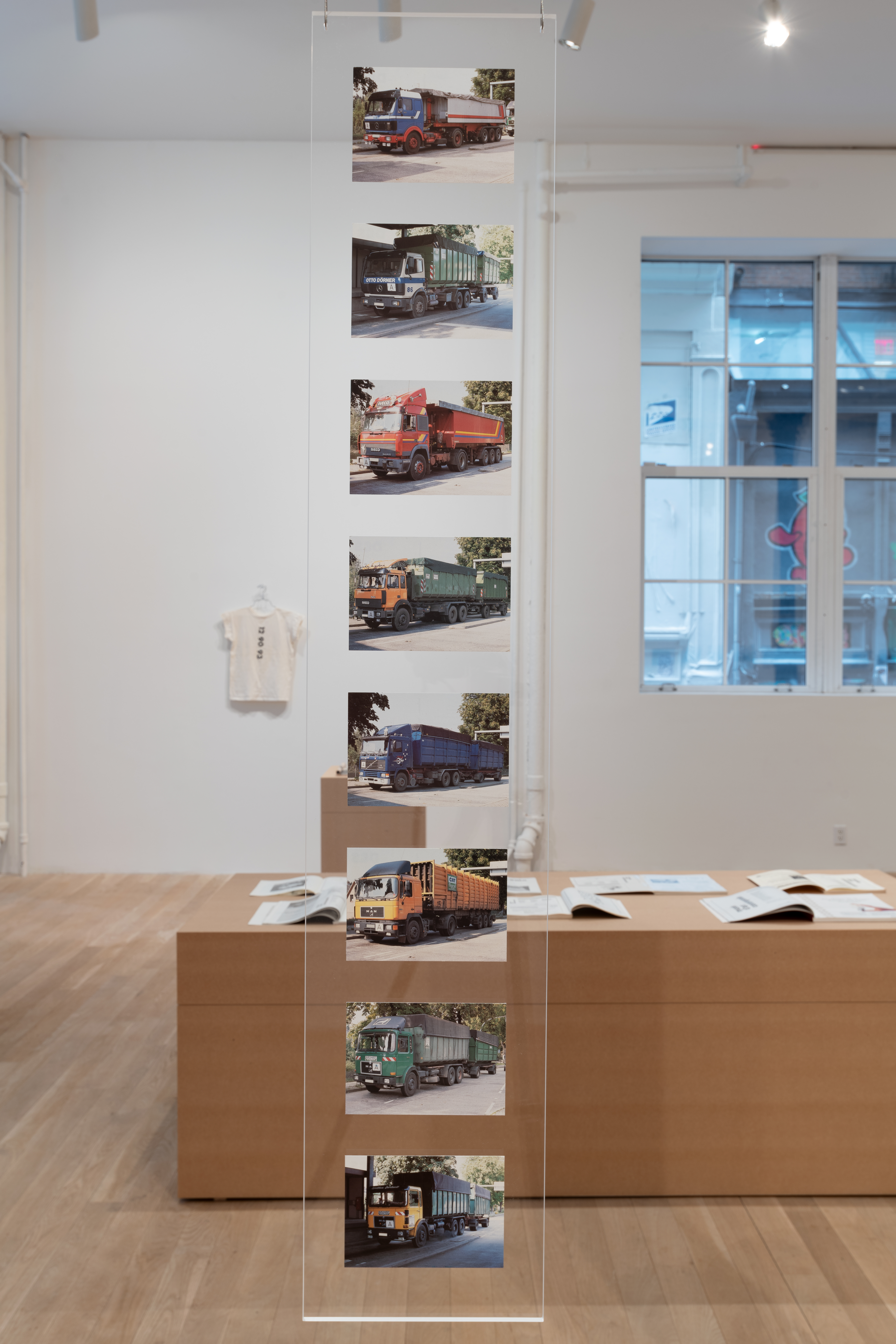 A long piece of plexiglass hangs vertically from the cieling in a gallery. Eight postcards are mounted onto the plexiglass. All of the postcards depict different garbage trucks, shown from the exact same angle. 