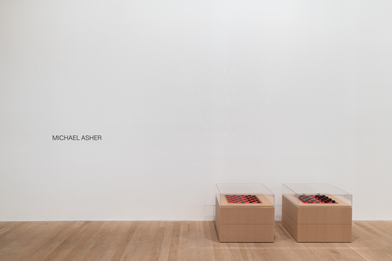 The text "MICHAEL ASHER" is handpainted in black onto a white gallery wall. To the right of the text are two identical plinths made of MDF. The plinths are low to the ground, and display checker board sets.