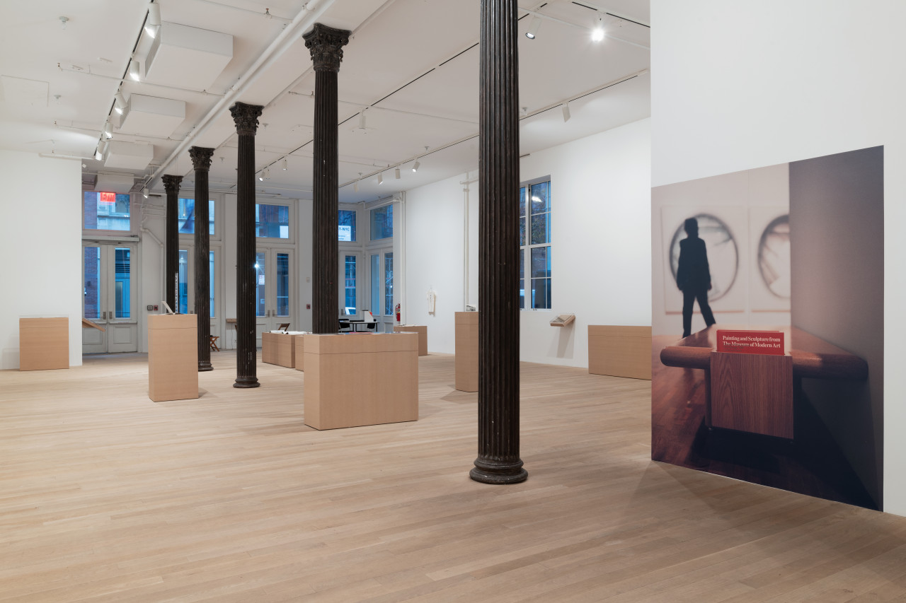 A large gallery space houses a variety of plinths made of MDF, atop of which are displayed archival objects such as journals and postcards. On the right wall in the foreground is a large photograph affixed to the wall which depicts a wooden museum bench with a box housing red deaccession catalogs made available within The Museum of Modern Art‘s exhibition space.