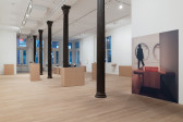 A large gallery space houses a variety of plinths made of MDF, atop of which are displayed archival objects such as journals and postcards. On the right wall in the foreground is a large photograph affixed to the wall which depicts a wooden museum bench with a box housing red deaccession catalogs made available within The Museum of Modern Art‘s exhibition space.
