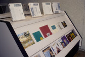Photograph of a white magazine stand with black trim. Along the top of the stand, four stacks of black-and-white journals rest on acrylic stands. A selection of art journals in a variety of shapes and colors sit along the stand's two rows.