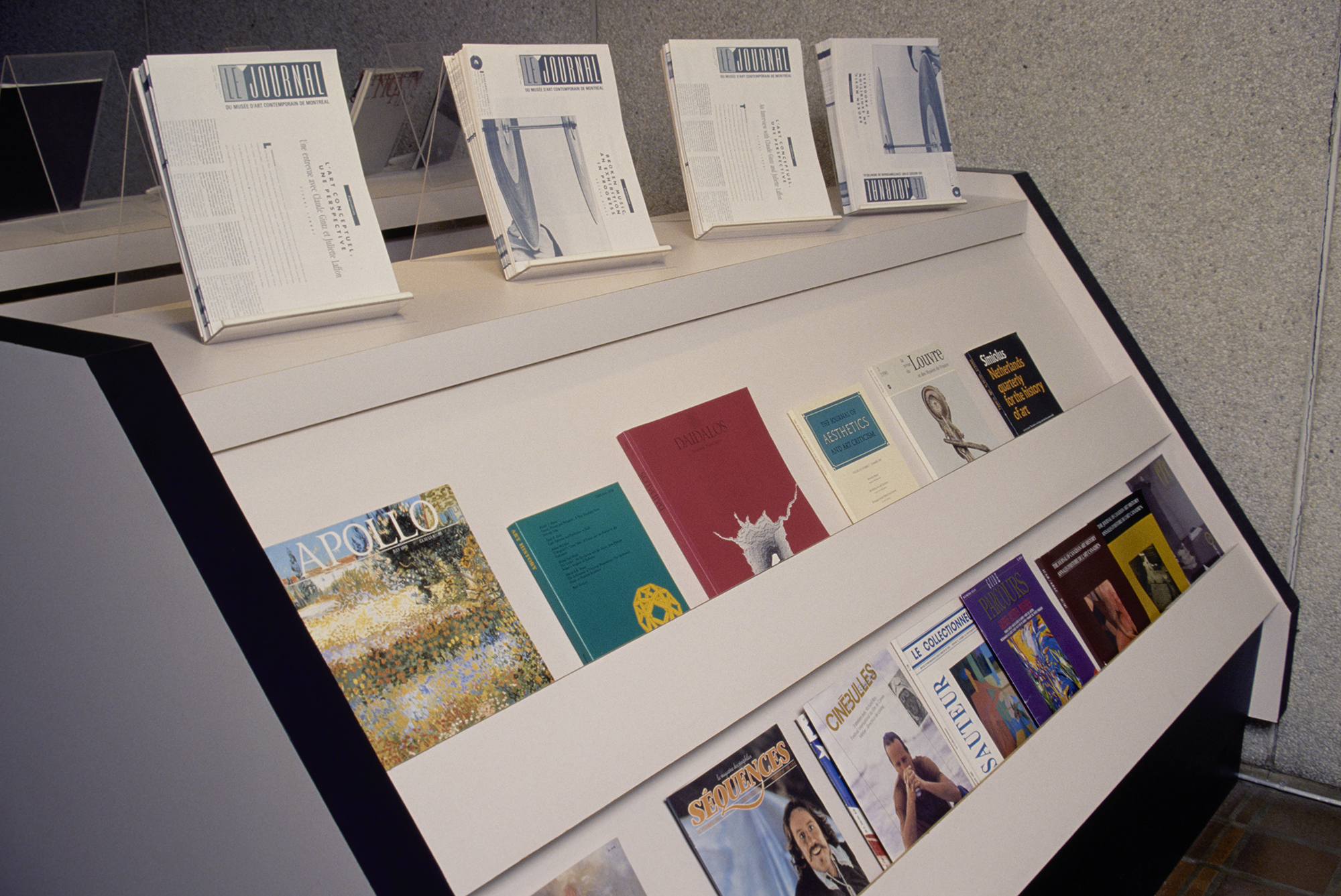 Photograph of a white magazine stand with black trim. Along the top of the stand, four stacks of black-and-white journals rest on acrylic stands. A selection of art journals in a variety of shapes and colors sit along the stand's two rows.