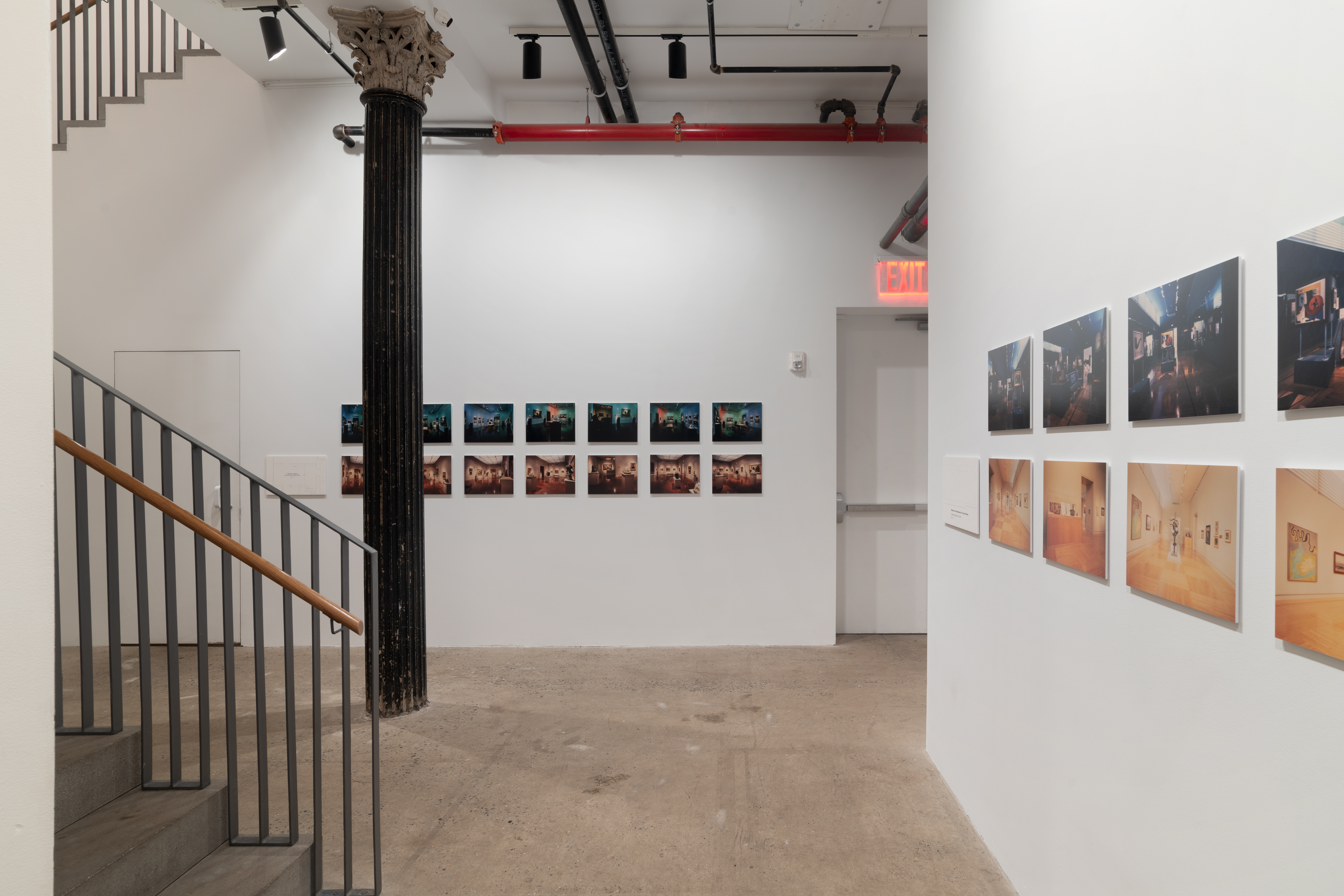 A set of stairs lead into a gallery space with two series of photographs on each wall. On the back wall a series of fourteen photographs are shown in two rows. Along the right wall, eight photographs in two rows are visible from this angle. In both series, the photographs on the top row are cool-toned and darker, and the photographs on the bottom row are much brighter and warm-toned. 