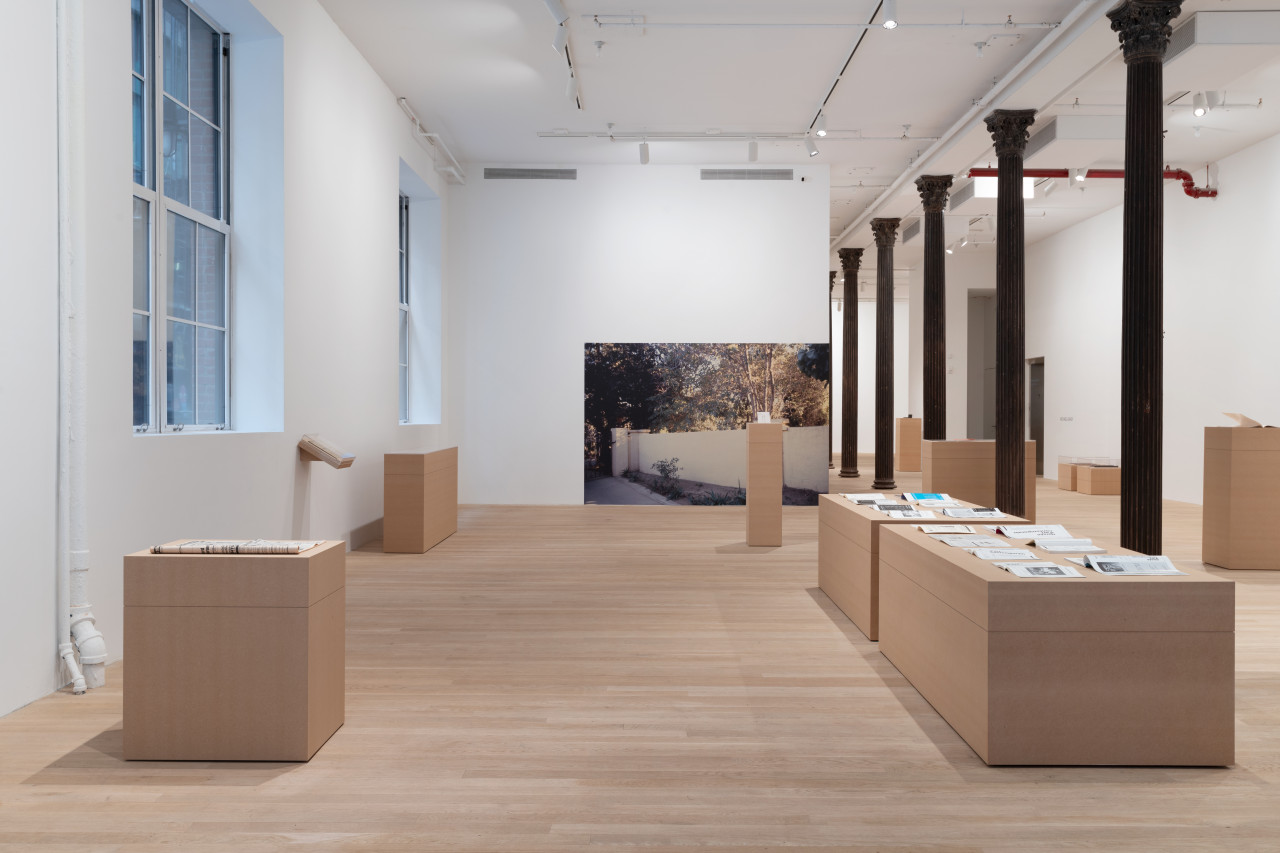 A large gallery space houses a variety of plinths made of MDF, atop of which are placed various archival objects. Along the far back wall is a large photograph affixed to the wall that depicts a driveway with a large white reconstructed wall.