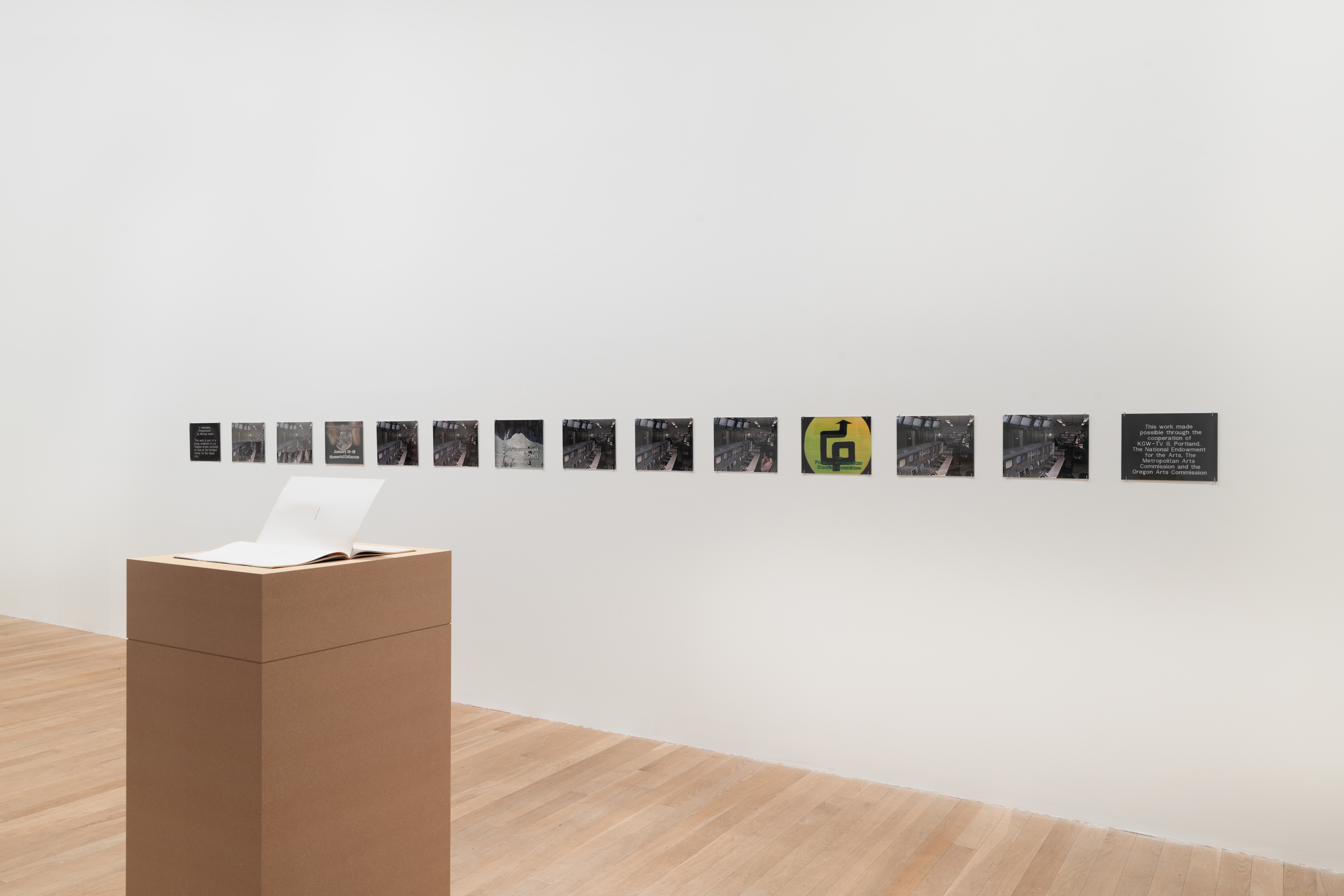 A series of reproduced photographic stills are displayed in a row along a white gallery wall. The stills are taken from a television broadcast and depict graphics from commercials, and the inside of a television studio. In the foreground is a magazine, displayed open on a plinth made from MDF.  