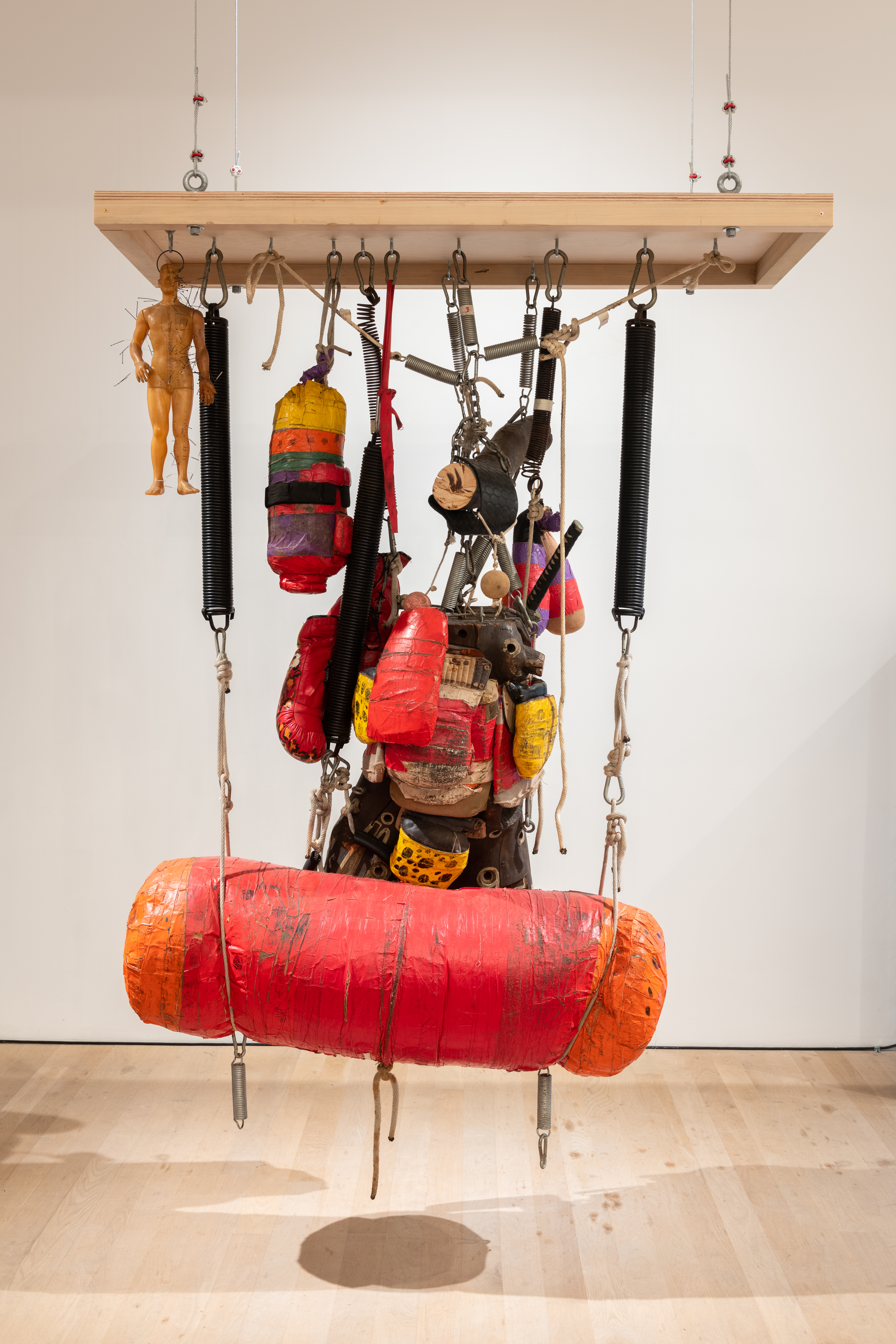 A color photograph of a red punching bag, other boxing equipment, and a human statue suspended from a rectangular wooden frame in a gallery space.