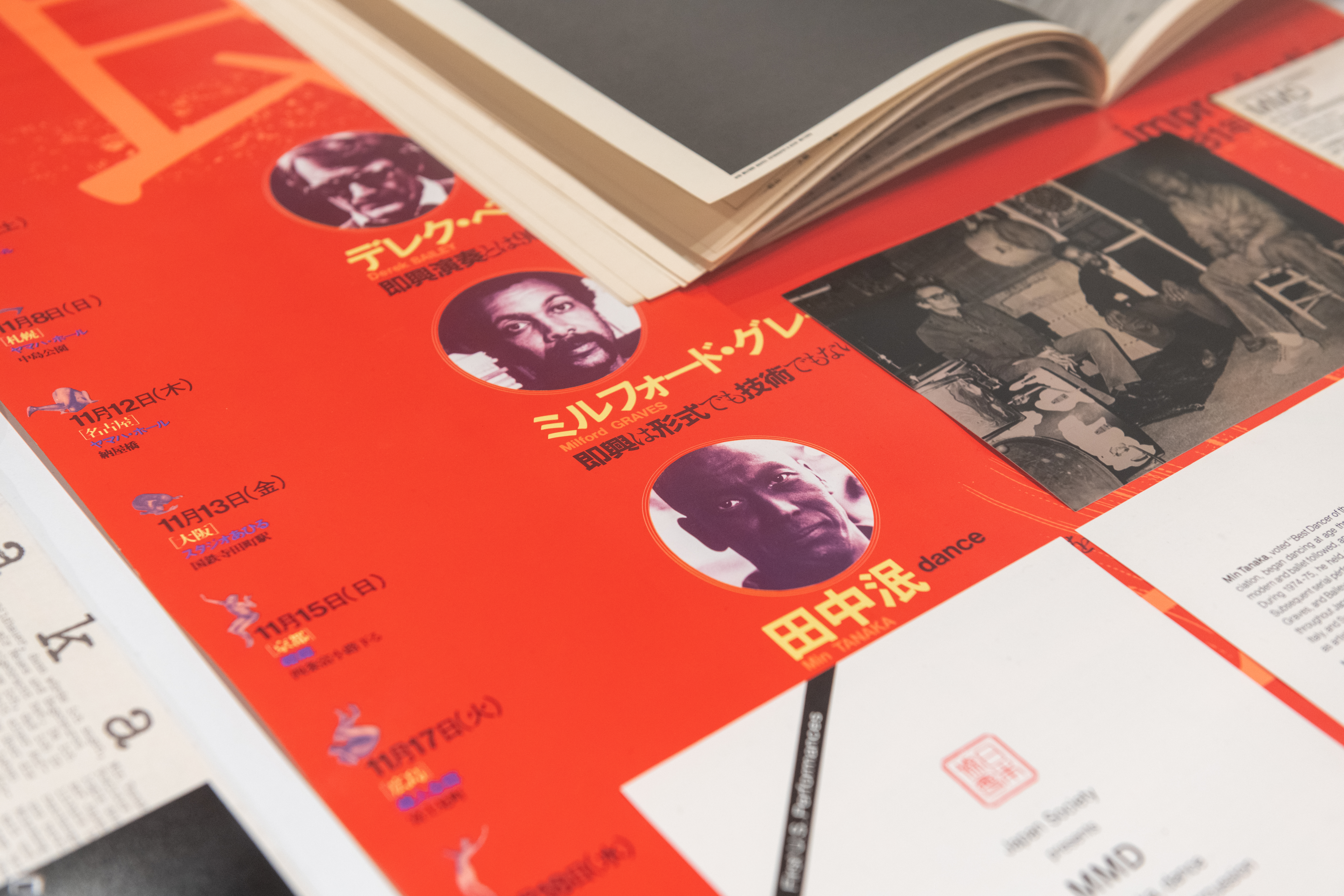 A color photograph of a red poster with three faces and Japanese captions. The poster is beneath other ephemeral material, including an open book, a pamphlet, and a black and white photograph.