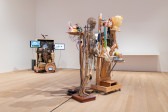 A color installation photograph of two sculptures in a gallery space. The sculpture closer to the camera is composed of a human skeleton, and anatomical models of human heads, a brain, and a spine. The sculpture in the background features several monitors, and is topped by a sculpture of an eagle in mid-flight.