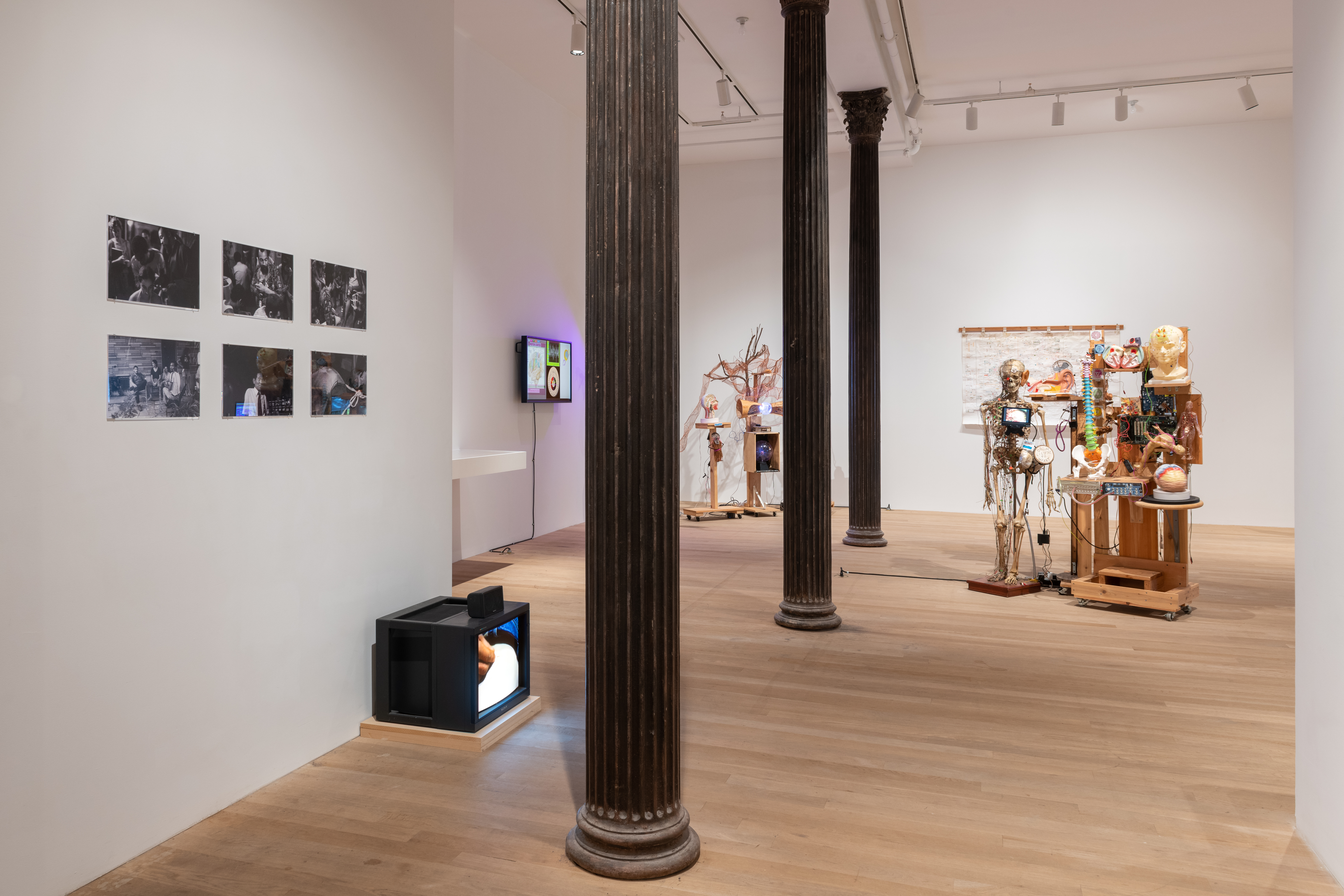 A color installation view of a gallery space. On the left wall are six black and white photographs and a television monitor positioned on the floor. In the background, another wall-mounted television monitor is visible along with a large wall hanging and two sculptures. The space is bisected by three dark columns.