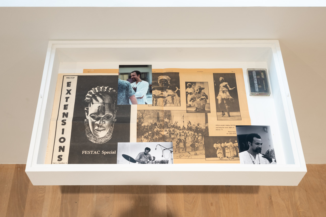 A color photograph of a white framed vitrine containing ephemera such as newsprint and several photographs.