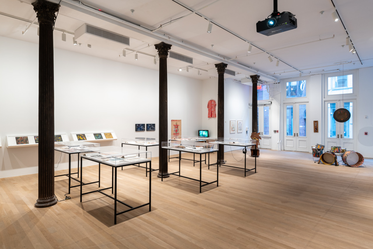 A color installation view of a gallery space with six glass vitrines positioned around four metal colums. A drum set and television monitor are also visible in the space.