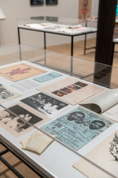 A color photograph of a vitrine containing ephemeral material, including pamphlets, photographs, and a blue flyer.