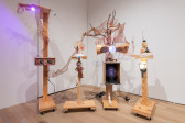 A color photograph of four sculptures mounted on wooden pedastals. The sculptures are connected by copper netting and are composed of objects including a model of a human head, a plasma globe, a drum, lights, and wires.