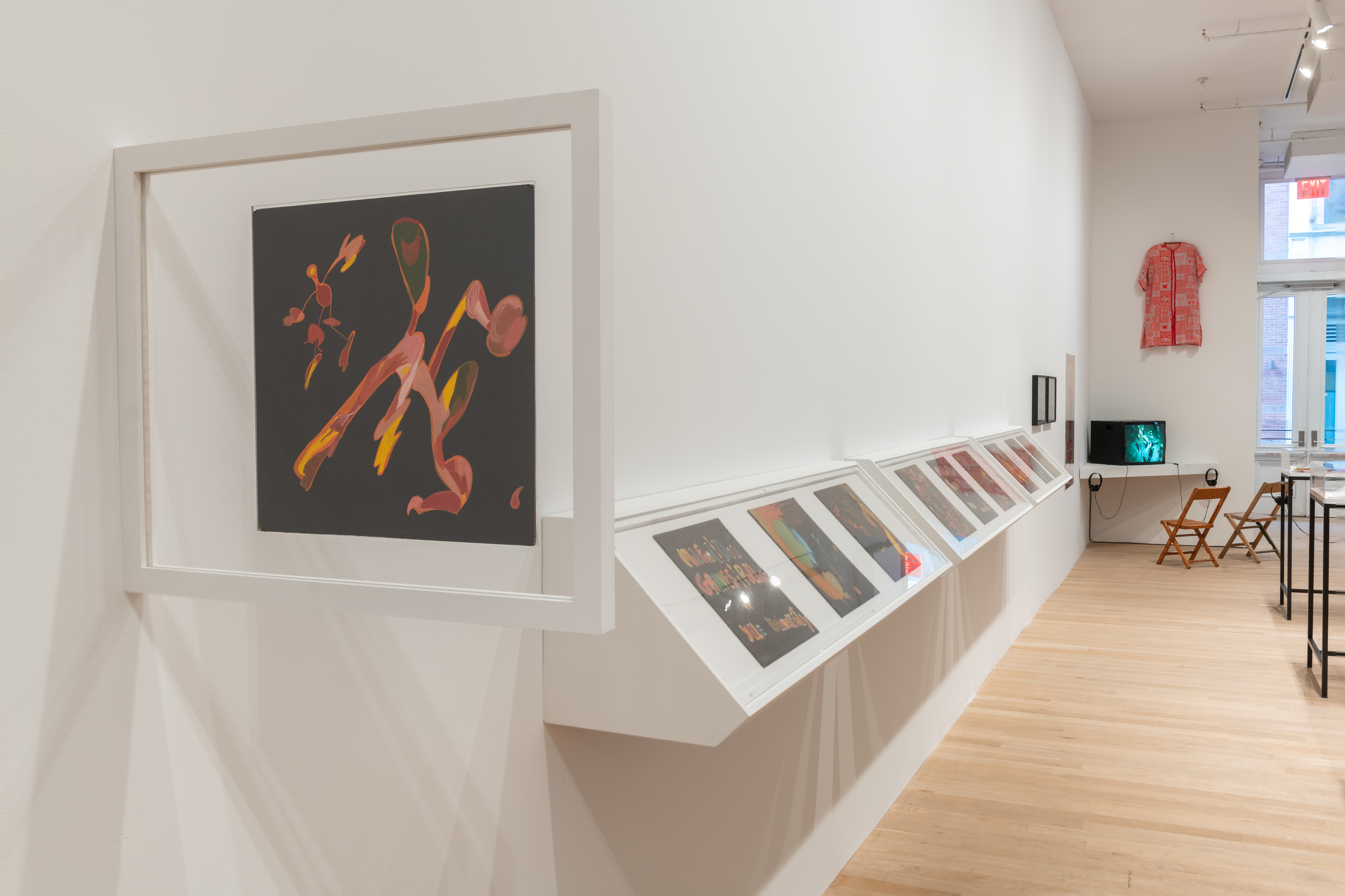 A color installation view of a gallery space. Several handpainted LPs line the left wall. A television monitor is visible in the back corner with two chairs in front. A red tunic hangs above the monitor.