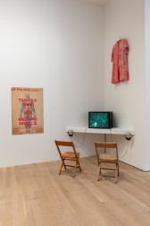 A color installation view of a gallery space. A poster with a drawing of a human body hangs on the left, a television monitor with two chairs is in the center of the image. Above the monitor, a red tunic hangs from the wall.