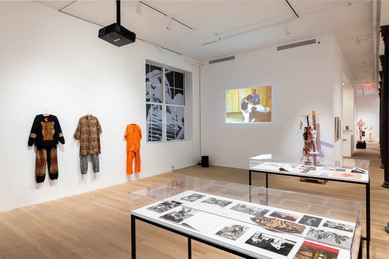 A color installation photograph of a gallery space. Three costumes hang from the left wall, a video is projected on a white wall in the background alongside a mixed-media sculpture. Two glass vitrines are visible in the foreground.