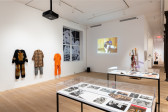 A color installation photograph of a gallery space. Three costumes hang from the left wall, a video is projected on a white wall in the background alongside a mixed-media sculpture. Two glass vitrines are visible in the foreground.