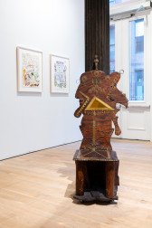 A color installation photograph of a gallery space, a painted wooden chair with an asymmetrical back is visible in the foreground. In the left background, two framed drawings hang from the wall.