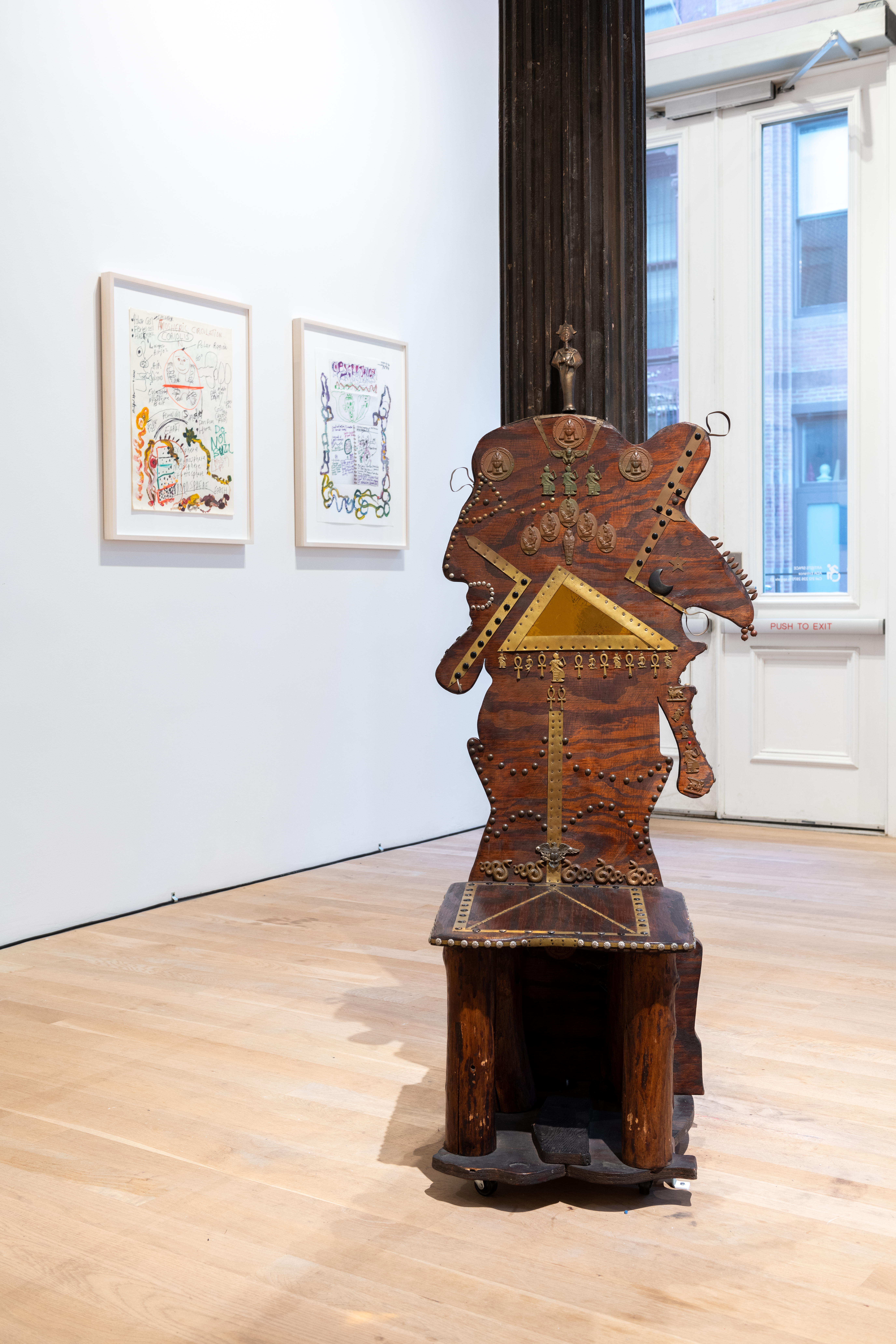 A color installation photograph of a gallery space, a painted wooden chair with an asymmetrical back is visible in the foreground. In the left background, two framed drawings hang from the wall.