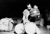 A black-and-white image of a man sitting on his knees with two African drums placed in front of him. His head looks downward while his left-hand hovers above the drums. His right hand firmly holds the side of a shaker instrument.