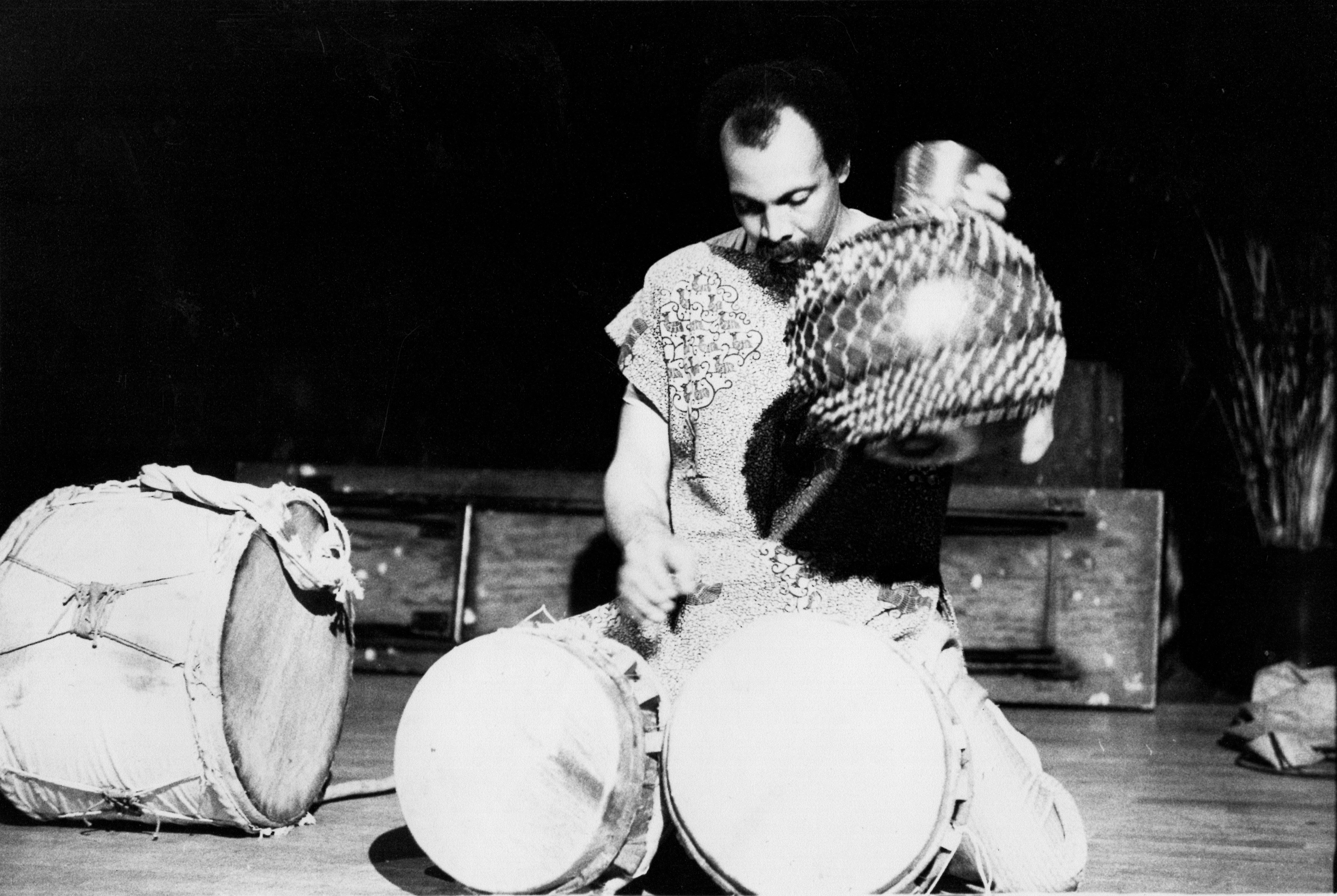 A black-and-white image of a man sitting on his knees with two African drums placed in front of him. His head looks downward while his left-hand hovers above the drums. His right hand firmly holds the side of a shaker instrument.