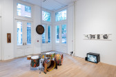 A color installation photograph of a gallery space with a painted drum set, a hanging gong. A television monitor is placed on the floor to the right with several LP covers hanging above.