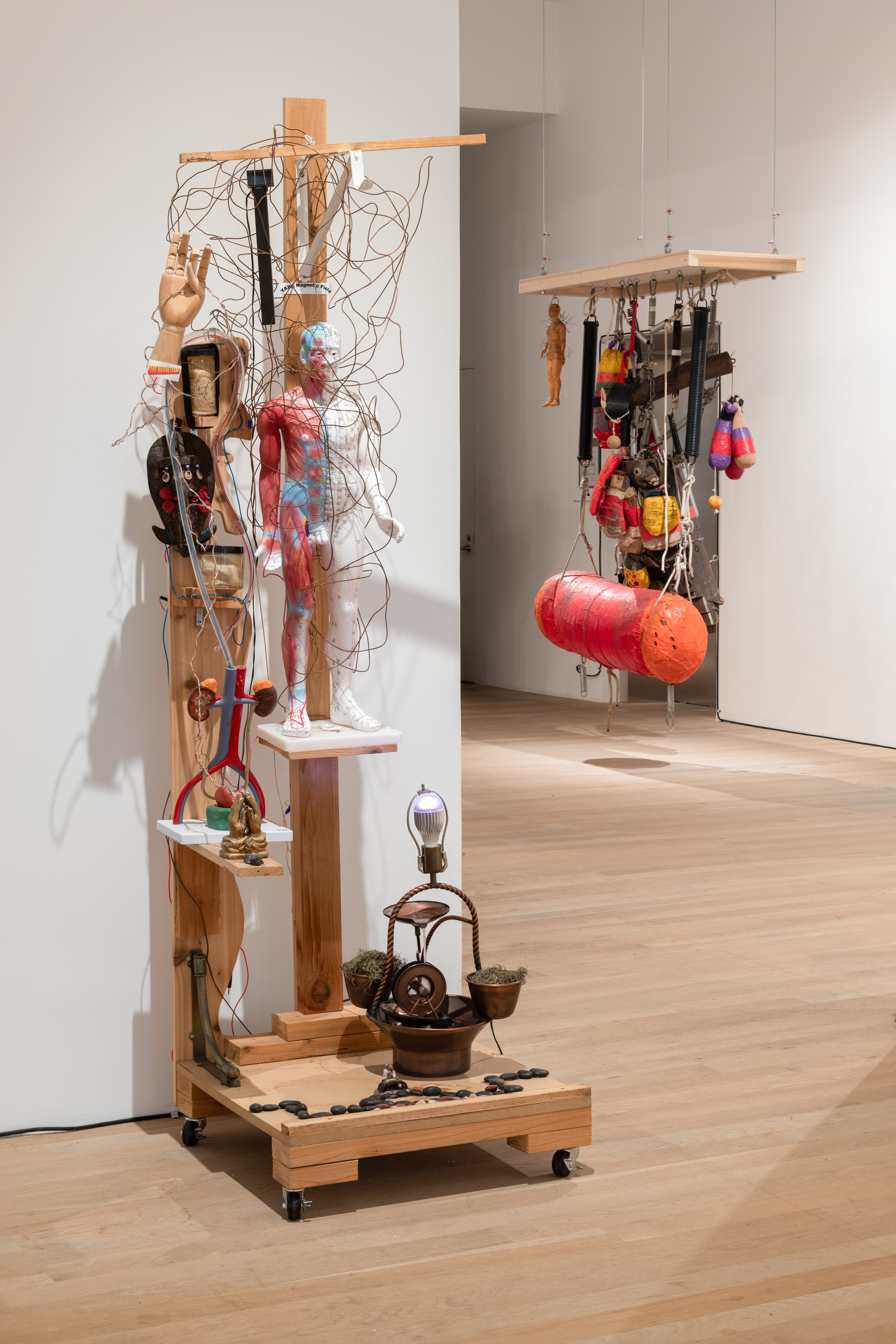 A color installation photograph of a gallery with two sculptures visible. The sculpture closer to the camera features an anatomical sculpture of a human body and hand, several wires, and a small water wheel. In the background, a punching bag and other boxing equipment are suspended from a rectangular wooden frame.