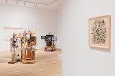 A color installation photograph of a gallery space. A collage hangs from the wall on the right, and two mixed-media sculptures are visible in the background along with a wall hanging.