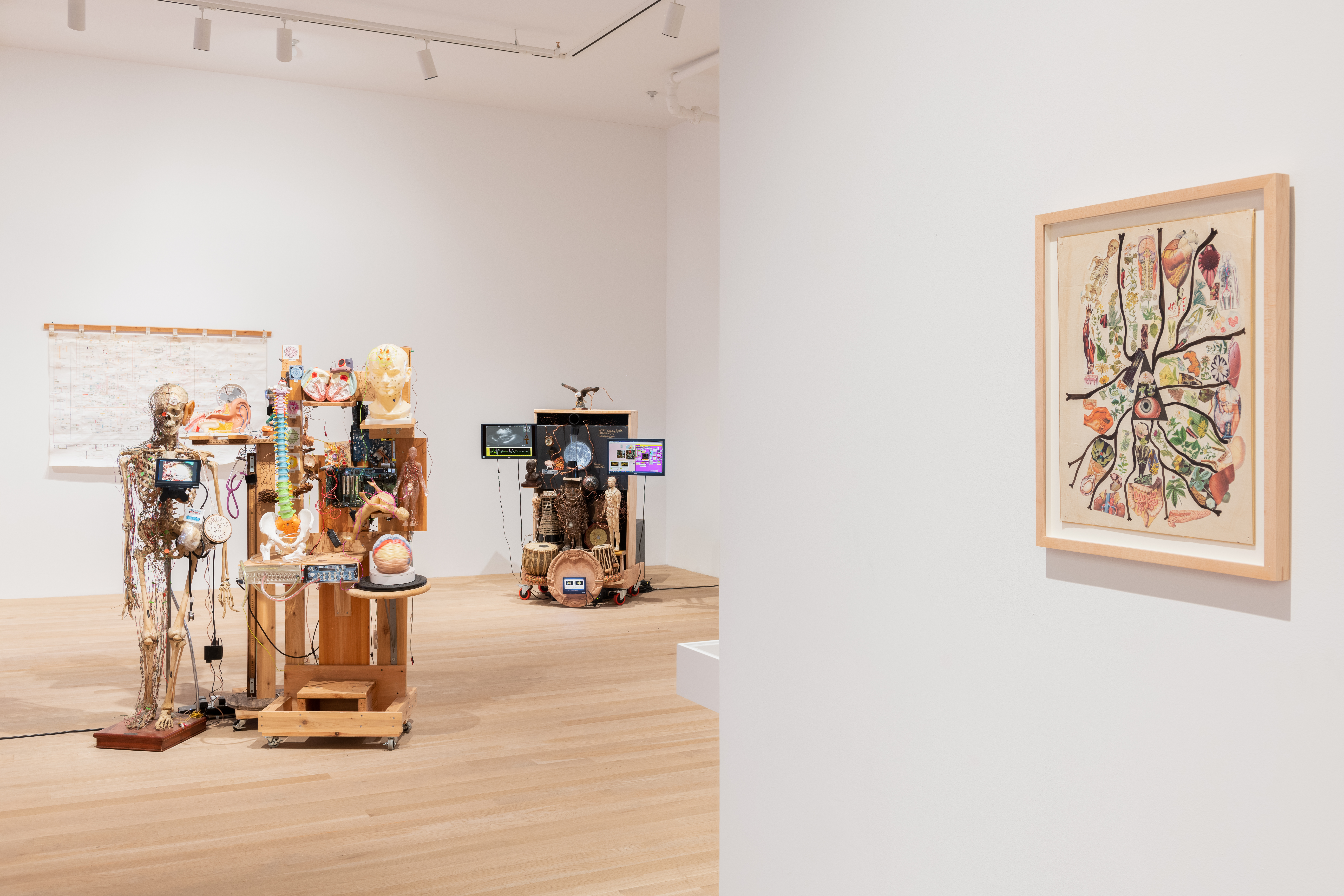 A color installation photograph of a gallery space. A collage hangs from the wall on the right, and two mixed-media sculptures are visible in the background along with a wall hanging.