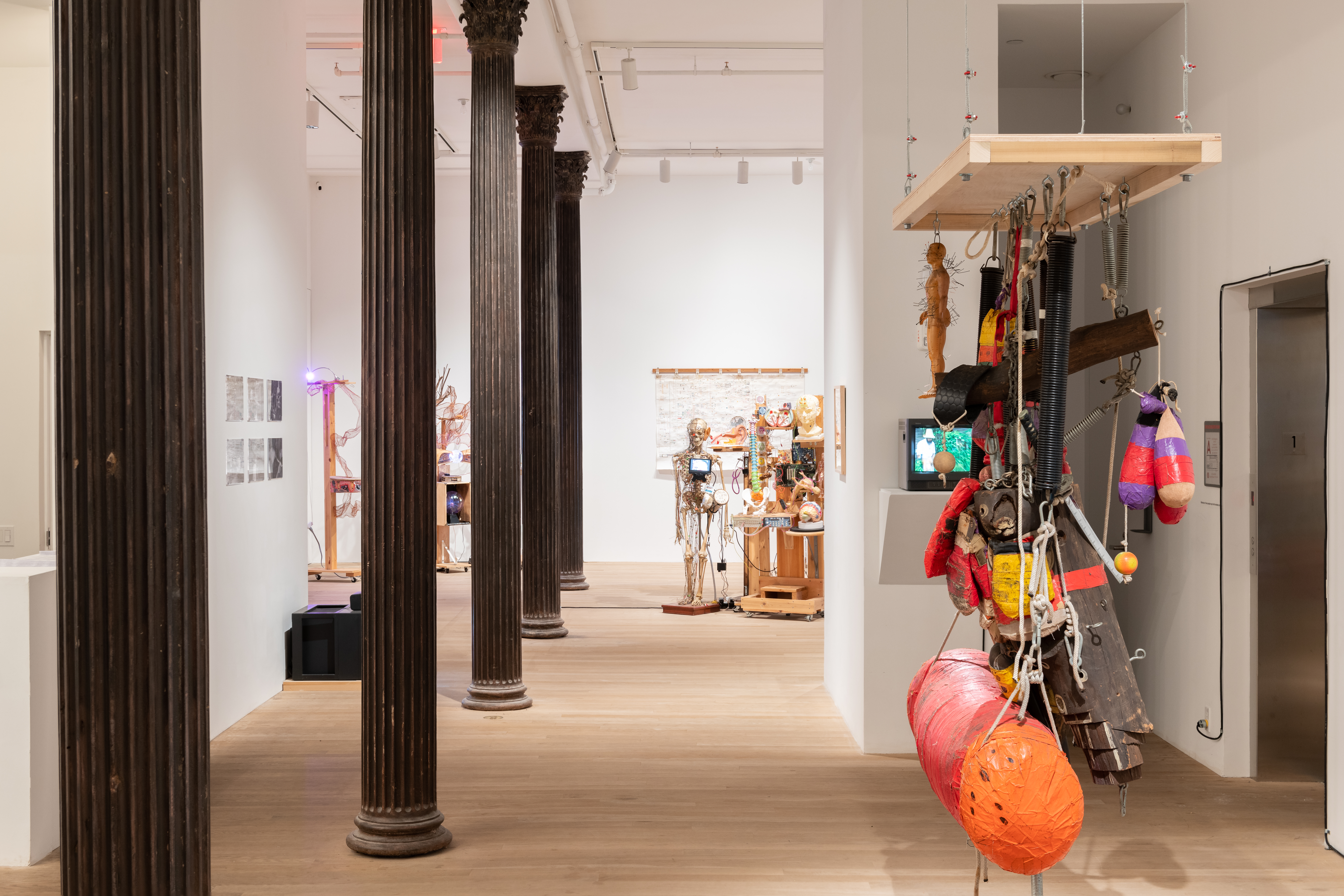 A color photograph of an installation in a gallery. Several dark metal columns line the left side of the space, and on the right a punching bag and other boxing equipment hang from a rectangular wooden frame. In the background, several more mixed-media sculptures are visible.