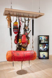 A color photograph of a a punching bag, other boxing equipment, and a sculpture of a human figure suspended from a rectangular wooden panel. In the background, three television monitors are stacked in a column, each playing a color video of figures in movement.