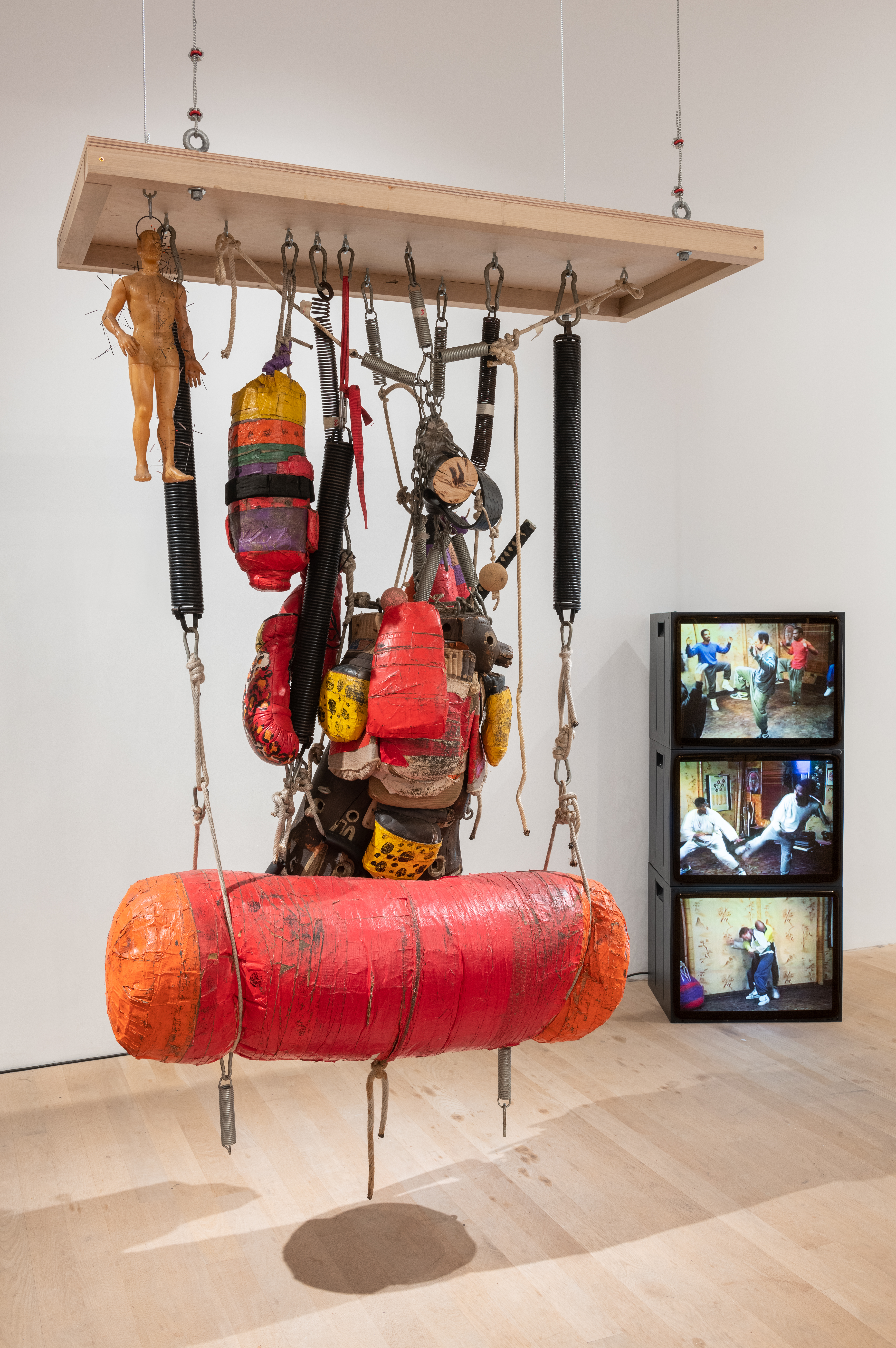 A color photograph of a a punching bag, other boxing equipment, and a sculpture of a human figure suspended from a rectangular wooden panel. In the background, three television monitors are stacked in a column, each playing a color video of figures in movement.