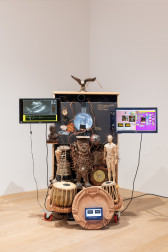 A color photograph of a sculpture composed of three monitors, drums, a human figure sculpture, and topped by a small sculpture of an eagle in mid-flight.