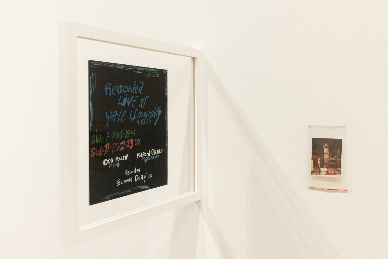 A color installation photograph of a hand painted LP in a glass frame and a small photograph in a plastic bag in a gallery space.