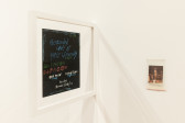 A color installation photograph of a hand painted LP in a glass frame and a small photograph in a plastic bag in a gallery space.