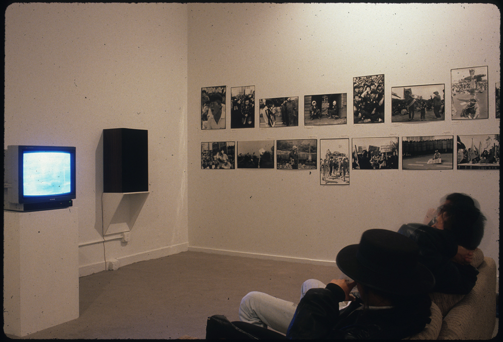 Several people seated on a couch watch a monitor set on a pedestal in front of them.  A large speaker is placed to the right of the screen, and on the wall alongside this are two rows of black and white photographs.