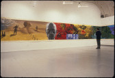 A photograph of a large, open gallery space.  The visible walls feature a continuous painting that wraps around the room, showing human figures on saturated blue, yellow, and red backgrounds.  A single person stands int he room and looks at the painting.