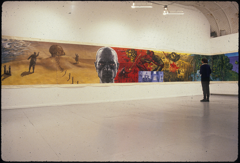 A photograph of a large, open gallery space.  The visible walls feature a continuous painting that wraps around the room, showing human figures on saturated blue, yellow, and red backgrounds.  A single person stands int he room and looks at the painting.