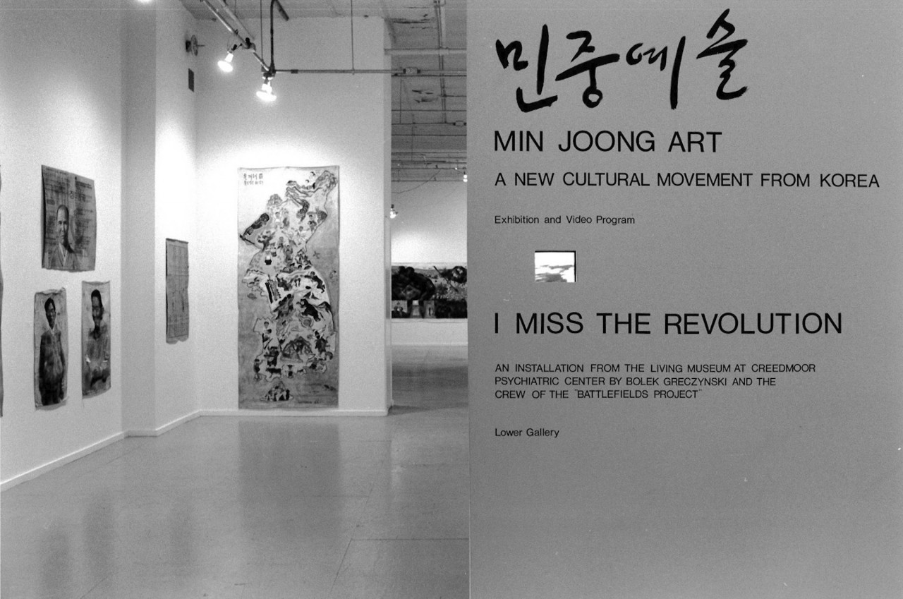A photograph of a gallery space.  The right half of the image is taken up by a wall with printed text identifying the exhibitions, "Min Joong Art: A New Cultural Movement from Korea," and, "I Miss the Revolution."