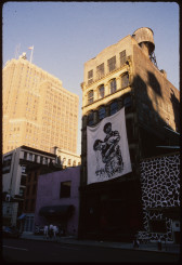 A photograph of building facades in Manhattan.  Hanging on the central facade is a piece of white fabric the length of two stories.  On the white fabric, there is a gestural painting of in a figure grapsing another figure from behind.  Below them is a line of text in Korean.