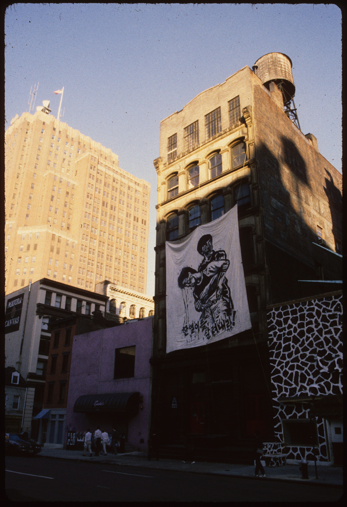 A photograph of building facades in Manhattan.  Hanging on the central facade is a piece of white fabric the length of two stories.  On the white fabric, there is a gestural painting of in a figure grapsing another figure from behind.  Below them is a line of text in Korean.