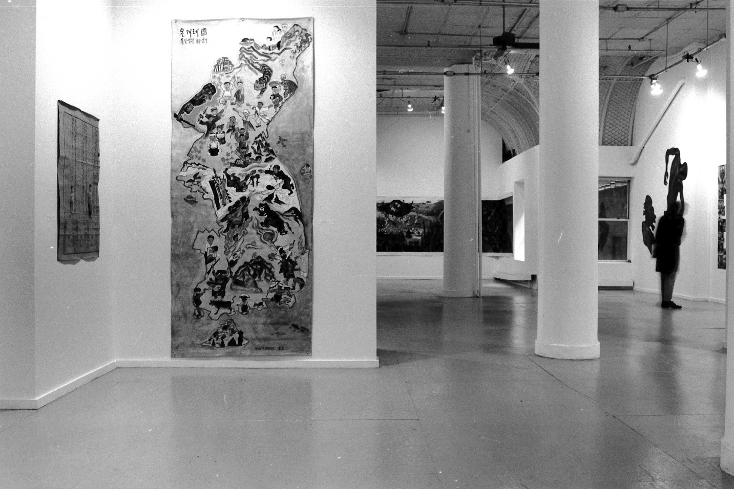 A photograph of a gallery space with white walls hung with artworks. In the foreground is a large painting that takes the form of a map of Korea.  In the background, another gallery with more artworks is visible.