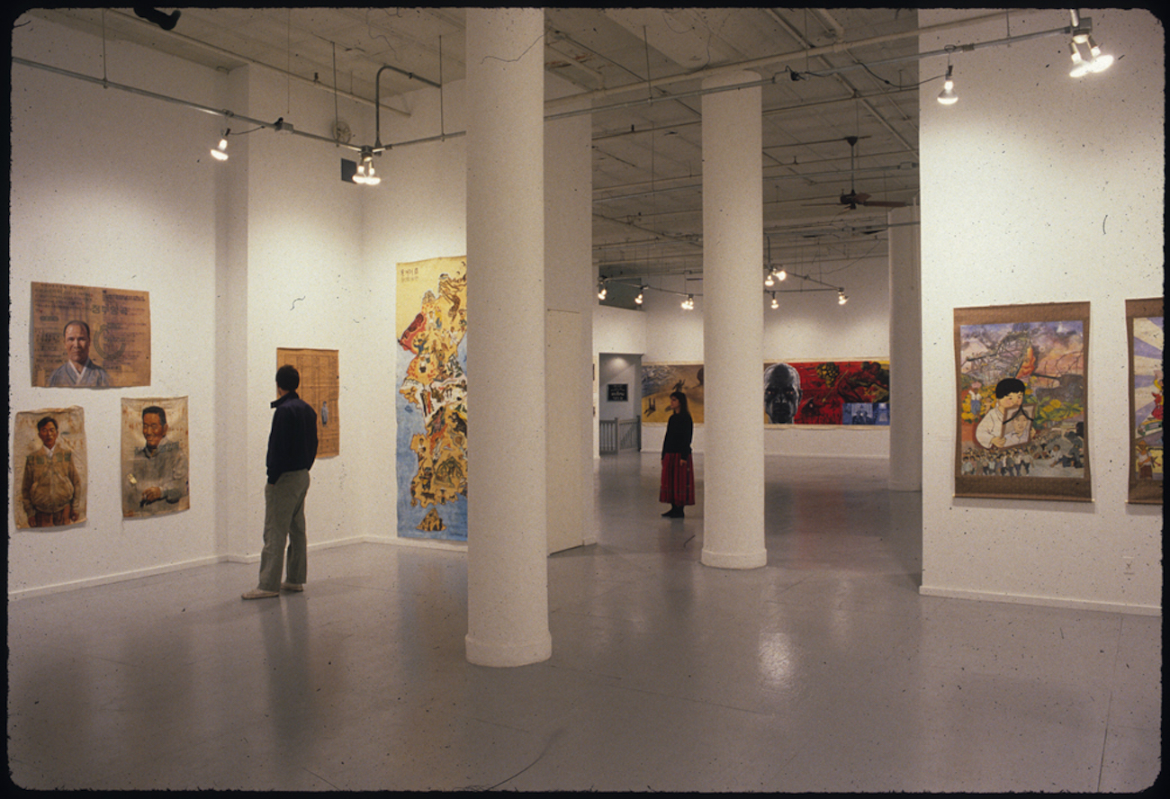 A photograph of a white walled gallery space densely hung with figurative paintings.