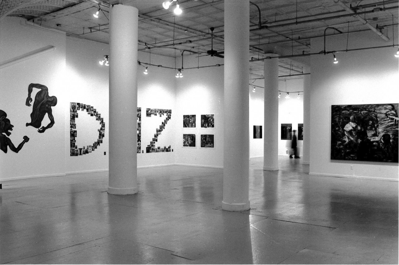 A photograph of a large gallery space with white walls and columns in the center of the room and artworks lining the walls.  In the center of the image, visible through the columns, are many small photographs hung in the shape of the letters "DMZ."